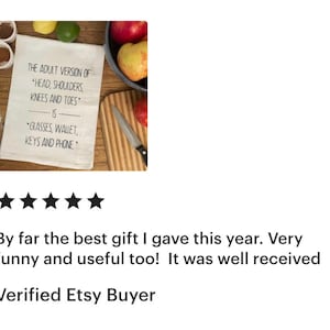 May include: A white rectangular kitchen towel featuring black printed text that reads: The adult version of head, shoulders, knees and toes is glasses, wallet, keys and phone. The towel is displayed alongside fresh fruit and a kitchen knife.