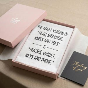 A beautiful pink gift box displaying a funny midlife screen printed humor towel that reads The adult version of head shoulders knees and towels is glasses wallet keys and phone"