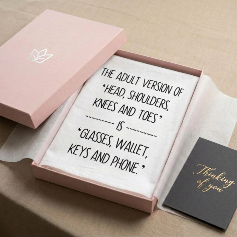 A beautiful pink gift box displaying a funny midlife screen printed humor towel that reads The adult version of head shoulders knees and towels is glasses wallet keys and phone"