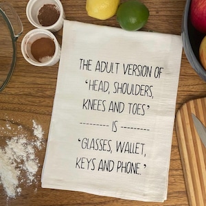 A funny white tea towel with black text that reads "THE ADULT VERSION OF 'HEAD, SHOULDERS, KNEES AND TOES' IS 'GLASSES, WALLET, KEYS AND PHONE.'" Towel is on a table surrounded by ingredients to make an apple pie. The Adult Version Of Head, Shoulders, Knees and Toes is Glasses, Wallet, Keys and Phone Kitchen Tea Towel Screen Printed