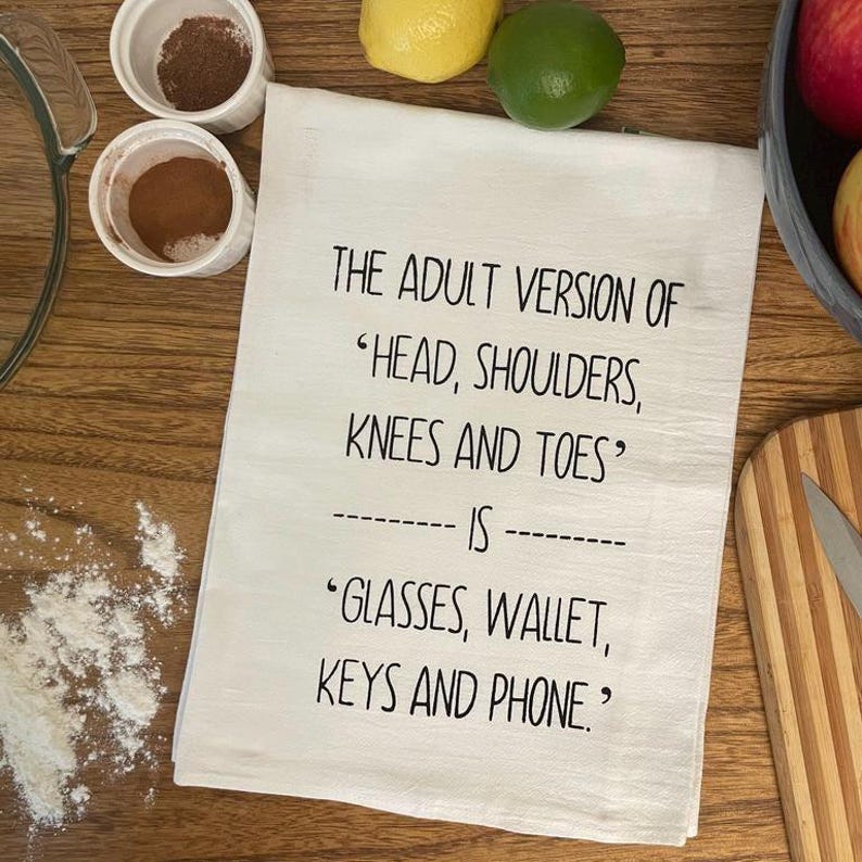 A funny white tea towel with black text that reads "THE ADULT VERSION OF 'HEAD, SHOULDERS, KNEES AND TOES' IS 'GLASSES, WALLET, KEYS AND PHONE.'" Towel is on a table surrounded by ingredients to make an apple pie. The Adult Version Of Head, Shoulders, Knees and Toes is Glasses, Wallet, Keys and Phone Kitchen Tea Towel Screen Printed
