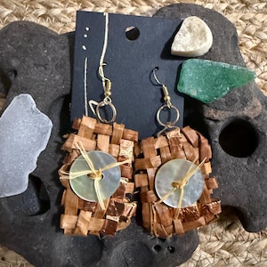May include: A pair of rectangular woven brown fiber earrings featuring gold-tone metal hooks and a central iridescent white circular button attached with light-colored raffia thread. The earrings are displayed on a black rectangular card.