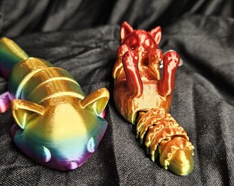 May include: Two 3D-printed fox figurines. One is a keychain with a rainbow gradient, the other is red and gold. The fox figurines are lying on a dark, textured surface. The rainbow fox is on its belly, while the red fox is on its back.