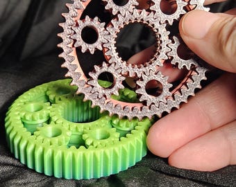 Op de afbeelding: Close-up van twee tandwielsets. Eén is groen, de andere is zilver en koperkleurig. Het zilveren tandwiel wordt in een hand gehouden. De tandwielen hebben veel tanden en worden waarschijnlijk gebruikt in een machine of mechanisch apparaat.