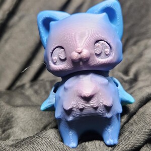 May include: A 3D-printed cat figurine in shades of blue and purple. The cat has large eyes, pointed ears, and a textured surface. The figurine is standing on a dark gray fabric.