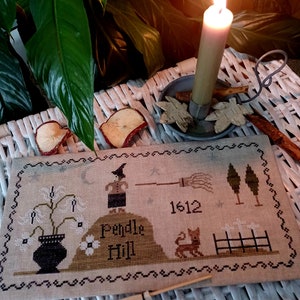 May include: A cross-stitch sampler featuring a witch on Pendle Hill, with the year 1612. The design includes a broom, trees, a cat, and flowers in a vase. The sampler is on a neutral fabric with a decorative border.