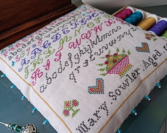 Könnte beinhalten: Ein weißes Leinenkissen mit bunter Kreuzstich-Stickerei des Alphabets, einem kleinen Blumenarrangement und einem Herzen. Das Kissen ist von blauen Perlen umgeben und der Text "Mary Sowler, Aged ..." ist in der rechten unteren Ecke gestickt.