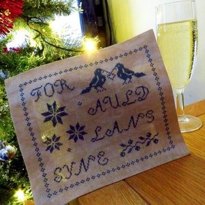 Puede incluir: Una tela beige con un diseño bordado en negro que presenta dos pájaros, copos de nieve y el texto "For Auld Lang Syne".