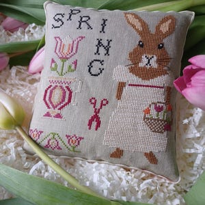 以下が含まれることがあります： 花かごを持ったウサギと「SPRING」の文字がクロスステッチで刺繍されたベージュのクッションです。