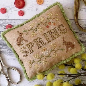 May include: A beige cross-stitch pillow with green trim features a floral design and the word "SPRING" in brown embroidery. The pillow is sitting on a white wooden surface with yellow flowers.