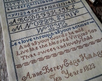 Könnte beinhalten: Ein Vintage-Sampler mit Kreuzstich-Buchstaben und -Zahlen in blauen und braunen Fäden auf cremefarbenem Stoff. Der Sampler enthält das Alphabet, Zahlen und ein Gedicht. Der Text lautet "Anne Berey Edge Hillage Ten Years 1823".