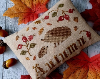 May include: A beige cushion with a cross-stitch design featuring two hedgehogs, a bush with red berries, and leaves. The cushion has the word "Autumn" stitched in brown thread.