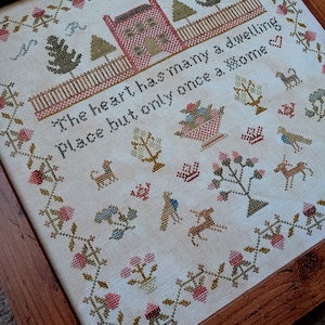 Peut inclure: Un sampler de point de croix encadré avec un fond blanc et une broderie colorée. Le sampler représente une maison, des arbres et le texte "The heart has many a dwelling place but only once a home."