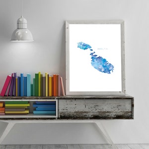 May include: A watercolor painting of the island of Malta in shades of blue. The painting is framed and hanging on a white wall above a bookshelf filled with colorful books.