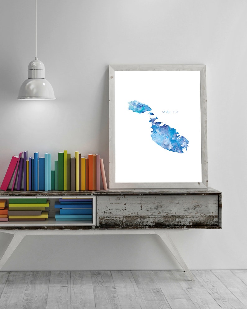 May include: A watercolor painting of the island of Malta in shades of blue. The painting is framed and hanging on a white wall above a bookshelf filled with colorful books.
