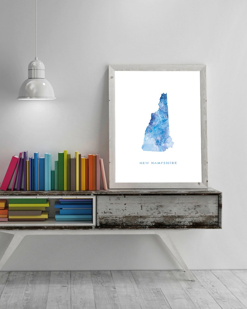 May include: A watercolor print of the state of New Hampshire in shades of blue. The print is framed and hanging on a white wall above a wooden shelf with colorful books.