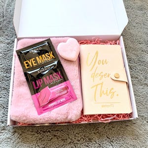 Op de afbeelding: Een roze cadeaudoos gevuld met zelfzorgartikelen. De doos bevat een roze pluche tasje met twee oogmaskers en een lipmasker, een roze hartvormige spons en een klein notitieboekje met de tekst "Je verdient dit." in gouden letters.