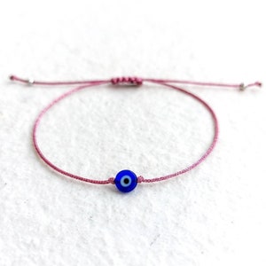 Puede incluir: Un brazalete de cuerda rosa con un amuleto azul de ojo turco. El amuleto tiene un centro blanco y un círculo azul alrededor.