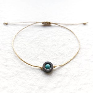 May include: A simple bracelet with a light brown cord and a round black bead with a turquoise stone in the center.