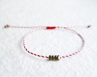 May include: A red and white string bracelet with gold beads. The bracelet is adjustable and can be worn on the wrist or ankle.