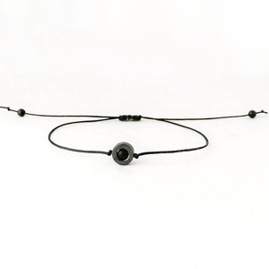 May include: A black string bracelet with a black bead in the center. The bead is a circle with a smaller circle inside.