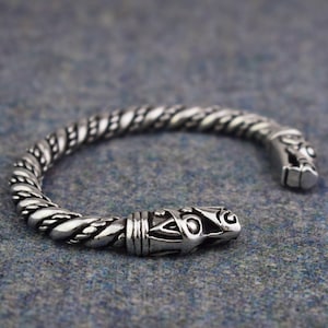 May include: A silver Viking-style cuff bracelet. The bracelet has a twisted rope design and animal head terminals. The open cuff design allows for easy wear. The bracelet is made of a shiny metal.
