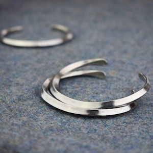 May include: Two silver metal bangles stacked on top of each other. The bangles are simple and have a smooth, polished finish.