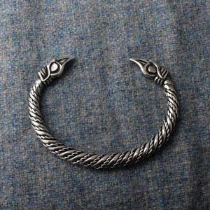 May include: A silver braided bangle with two bird head ends. The bangle is on a grey textured surface.