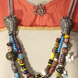 May include: A colourful, multi-strand beaded necklace with silver-coloured metal end caps. The beads are in shades of blue, yellow, red, and brown. A silver-coloured brooch is pinned to the garment. The necklace is a statement piece.