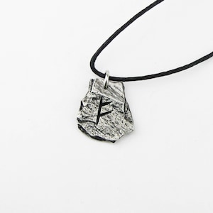 May include: A silver pendant necklace with a black cord. The pendant is shaped like a rock with a rune carved into it. The rune is the Elder Futhark rune for 'Fehu', which represents wealth, prosperity, and abundance.