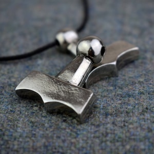 May include: A silver-colored pendant in the shape of Thor's hammer, Mjolnir, hangs from a black cord. The hammer has a detailed design and is shown in a close-up shot. The background is a textured, blue-gray surface.