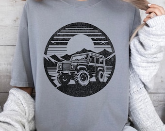 May include: A grey crew neck t-shirt featuring a black circular graphic print. The design depicts an off-road vehicle positioned in front of a mountain range and a sun, rendered in a distressed, vintage-inspired style with horizontal line details.