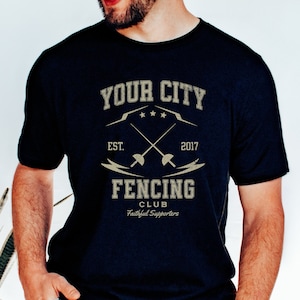 May include: A navy blue t-shirt with a vintage-style graphic design. The design features the text "YOUR CITY" with three stars, "EST. 2017", crossed fencing swords, and the text "FENCING CLUB Faithful Supporters".