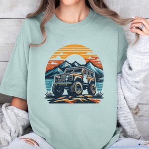 May include: A light blue t-shirt featuring a vintage-style graphic of a jeep driving through a mountain landscape with a sunset in the background.