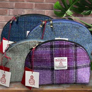 May include: Four Harris Tweed cosmetic bags in shades of blue, gray, and purple. Each bag has a zipper closure and a tag with a logo. The bags are made of a textured fabric with a plaid or herringbone pattern.