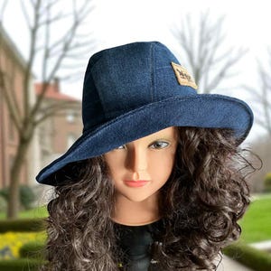 May include: A dark blue denim hat with a wide brim. The hat features a small, rectangular patch with a logo on the front. The hat is displayed on a mannequin head with long, curly brown hair.