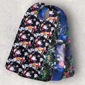 May include: A set of four layered bibs featuring distinct patterns: one with a black background and stylized cartoon faces, one with a dark green floral print, one with a blue space theme showing planets, and one with a colorful nebula galaxy design.