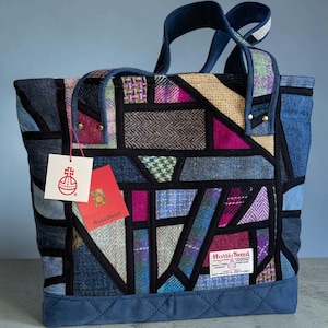 May include: A tote bag constructed from a patchwork of multicoloured Harris Tweed wool fabrics and blue denim, featuring black fabric borders, two top handles, and a quilted blue base. Two product tags are attached, one white and one red.