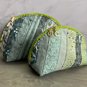 May include: Two semi-circular zipped pouches featuring a patchwork design of various green and grey fabrics. The patterns include floral, leaf, and striped motifs. Each pouch has a lime green zip trim and gold-toned circular zip pulls.