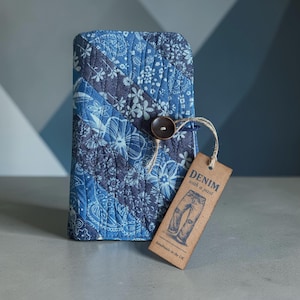 May include: A quilted blue denim wallet with a button closure. The wallet features a patchwork design with floral and paisley patterns. A brown tag with the text "DENIM with a past" and an illustration of jeans is attached.