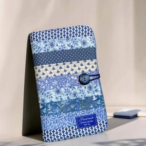 May include: A rectangular fabric case featuring horizontal patchwork stripes in various shades of blue and white, including floral, polka dot, and geometric patterns. A dark button closure is attached with an elastic loop. A label reads Hand made SPECIAL EDITION FOR YOU.