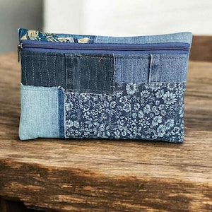 May include: A rectangular denim pouch with a blue zip closure. The pouch is made of various denim patches in different shades of blue, including a floral patterned fabric. The pouch is approximately 20 cm wide and 13 cm tall.