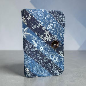 Op de afbeelding: Een blauwe stoffen portemonnee met een patchwork ontwerp. De portemonnee heeft verschillende tinten blauw met bloemen- en paisley patronen. Het heeft een knoopsluiting met een donkerbruine knoop en een blauwe elastische lus.