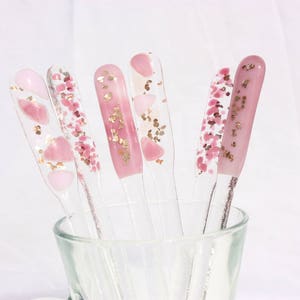 May include: A collection of clear nail art display sticks, each adorned with pink and gold glitter and heart-shaped designs. The sticks are arranged in a clear glass, showcasing the variety of decorative nail art options.