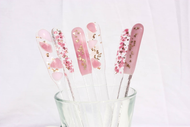 May include: A collection of clear nail art display sticks, each adorned with pink and gold glitter and heart-shaped designs. The sticks are arranged in a clear glass, showcasing the variety of decorative nail art options.