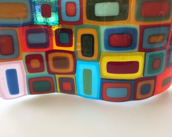 May include: A colorful glass art piece with a wavy, abstract design. The piece is made up of many small squares of different colors, including red, yellow, blue, green, and brown.