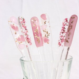 May include: A collection of clear, decorative stir sticks with pink and gold accents. The stir sticks are in a clear glass and feature various designs, including pink hearts and gold flakes. The overall aesthetic is delicate and feminine.