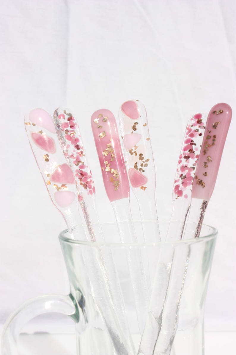 May include: A collection of clear, decorative stir sticks with pink and gold accents. The stir sticks are in a clear glass and feature various designs, including pink hearts and gold flakes. The overall aesthetic is delicate and feminine.