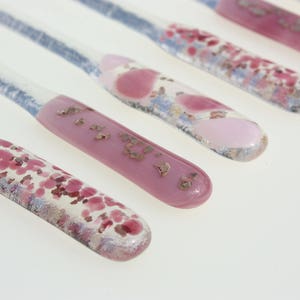 May include: Close-up of six glass stir sticks with decorative ends. The ends feature pink, red, and gold accents. The clear stems have a blue hue. The sticks are arranged diagonally against a white background.