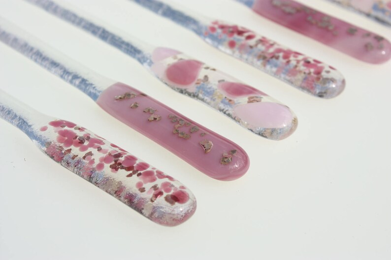 May include: Close-up of six glass stir sticks with decorative ends. The ends feature pink, red, and gold accents. The clear stems have a blue hue. The sticks are arranged diagonally against a white background.